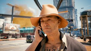 Nahaufnahme eines telefonierenden Menschen mit Strohhut, im Hintergrund die Stadt Night City. Screenshot aus demSpiel Cyberpunk 2077