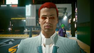 Portät eines Mannes in kurzen, roten Haaren, kleiner Brille mit roten Gläsern und teurem Anzug auf der Strasse von Night City. Screenshot aus dem Spiel Cyberpunk2077
